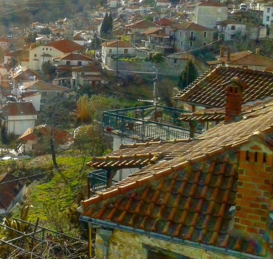 Rapsani in morning light: tiled-roof houses climbing a green slope, a roof with a brick chimney in front